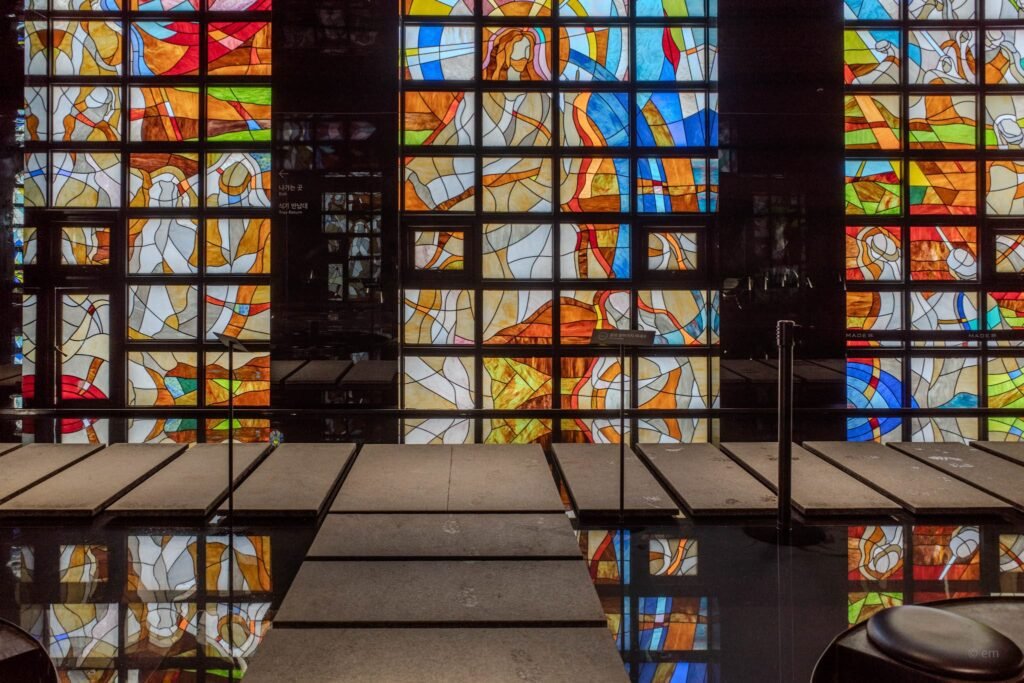 Colorful stained-glass wall inside a church-like interior on Yeongjong-do, with vibrant geometric panels, glossy black surfaces, and the MADE林 forest-inspired design.